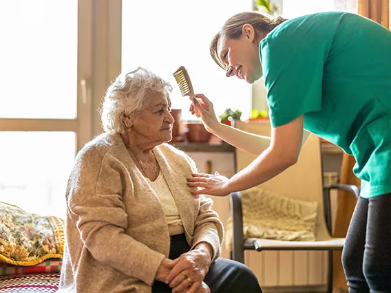 A female nurse aid brushes the hair of an elderly woman sitting on a bed