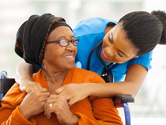 An African-American woman in a wheelchair being hugged from behind by a care worker.