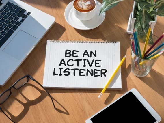 A notebook on a desk with the words "Be an active listener" written on it, surrounded by a laptop, coffee cup, tablet, glasses, and colored pencils.