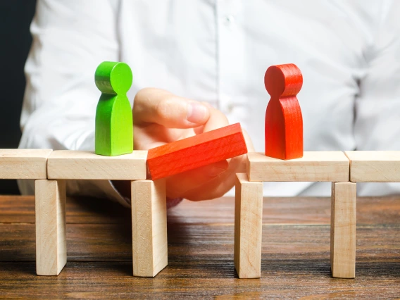 A person places a red block to bridge a gap between two wooden figures, symbolizing connection or resolving conflict in difficult conversations.