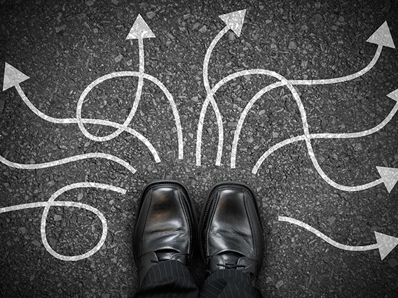 Black dress shoes standing above multiple white arrows pointing in different directions on asphalt, representing decision-making and choosing direction in life.