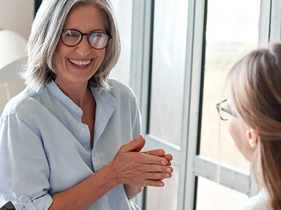 Two professional women having friendly conversation in bright office setting, one holding coffee cup while seated at desk with colleague standing nearby