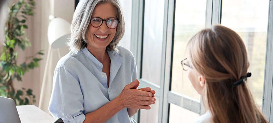 Two professional women having friendly conversation in bright office setting, one holding coffee cup while seated at desk with colleague standing nearby