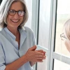 Two professional women having friendly conversation in bright office setting, one holding coffee cup while seated at desk with colleague standing nearby